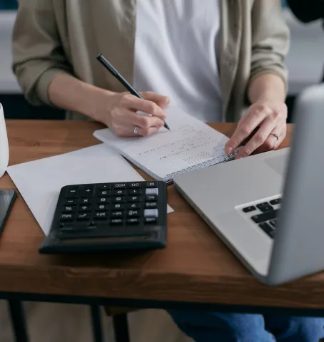 Hands writing in a notebook beside a calculator and laptop.